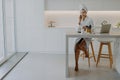 Pleased young woman dressed in bathrobe poses bare feet at chair in spacious kitchen sits at table with laptop and cosmetic Royalty Free Stock Photo