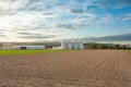 plowed field with silver silos and a farmhouse at the horizon