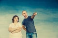 A plump red-haired attractive woman and a tall bald man on a blue background, look to the side, he points a finger,