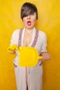 Plump woman with short black hair stands in a pink Bathrobe with a rubber cleaning glove on her hand and a dustpan on a yellow Stu