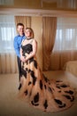 A plump woman in royal dress and a skinny man together in a honeymoon hotel room. An adult newlywed couple embraces in