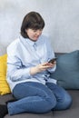 Plump young woman in strict shirt sits on couch with phone in hands. Vertical frame