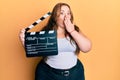 Plus size caucasian young woman holding video film clapboard covering mouth with hand, shocked and afraid for mistake