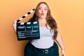 Plus size caucasian young woman holding video film clapboard scared and amazed with open mouth for surprise, disbelief face