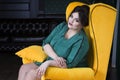 Plus size fashion model in green evening dress sitting in yellow armchair, fat woman on luxury interior