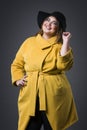 Plus size fashion model in yellow coat and black hat, fat woman on gray background, overweight female body