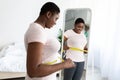 Plus size black woman measuring waist with tape, standing near mirror, satisfied with results of slimming diet at home Royalty Free Stock Photo