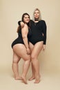 Plus Size Models. Full-figured Women Full-length Portrait. Brunette And Blonde In Black Bodysuits Posing On Beige Background.