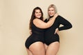 Plus Size Models. Full-figured Women Portrait. Brunette And Blonde In Black Bodysuits Posing On Beige Background.