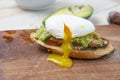 Poached egg on a slice of toast bread, with guacamole and dripping yolk, on a wooden board