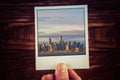Polaroid postcard photograph of Melbourne skyline from waterfront held by male hand over wooden table with copy space. Travel