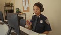 Policewoman in uniform working at a desk with computer and headset in a police station, taking notes, showcasing a professional Royalty Free Stock Photo