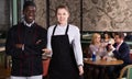 Polite waitress standing in restaurant hall with cheerful African American chef, meeting guests Royalty Free Stock Photo