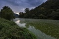 A polluted river flowing between trees and bushes under dark clouds at sunset Royalty Free Stock Photo