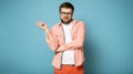 Pompous bearded man with glasses, shows a hand gesture to the side and looks at the camera. Isolated on a blue