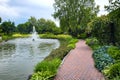 A pond filled with water with a spray jet fountain in a park with pedestrian sidewalks.