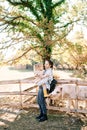 Pony sniffs the boot of a little girl sitting in her mother arms near a wooden fence in a pasture