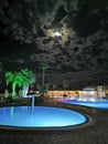 Pools of water illuminated with blue light surrounded by palm trees against a dark sky with a moon and beautiful clouds Royalty Free Stock Photo