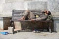 Poor homeless beggar man or refugee sleeping on a dirty wooden bench in a one-way street in the city during day time. Social