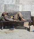 Poor homeless beggar man or refugee sleeping on a dirty wooden bench in a one-way street in the city during day time. Social