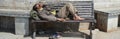 Poor homeless beggar man or refugee sleeping on a dirty wooden bench in a one-way street in the city during day time. Social