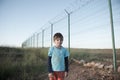 Poor little caucasian refugee boy in dirty clothes standing near high fence with barbed razor wire at state border