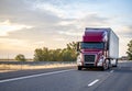 Popular model burgundy big rig semi truck transporting commercial cargo in dry van semi trailer running on the evening highway Royalty Free Stock Photo