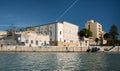 At the port of Mazara del Vallo, houses line the river against a blue sky. A house is derelict and without a roof. Behind other