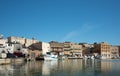 At the port of Mazara del Vallo there are many houses on the river against a blue sky. The houses are reflected in the water. Old