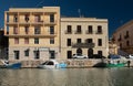 At the port of Mazara del Vallo there are two houses by the river against a blue sky. The houses have many windows and balconies.