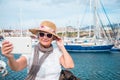 A port with yachts and sailboats. An elderly woman with gray hairs takes a selfie with the scenery with her tablet. Cloudy and