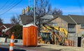 Portable bathroom at construction site at intersectio of urban roads with backhoe in background and traffic cones around