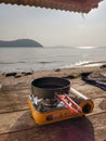 Portable tourist gas stove and cooking pan on wooden table at sandy beach with morning sunlight. Camping cookware outdoors.