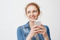 Portait of cute tender redhead teenage girl with messy hair, looking aside and smiling while holding smartphone and