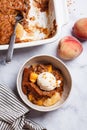 Portion of peach cobbler with scoop of ice cream in bowl, white marble background