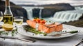 portion of smoked salmon steak and asparagus in a white plate on a white tablecloth in front of Icelandic waterfalls