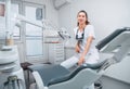 Portraait of sincerely smiling young dentist woman dressed white medical scrubs uniform sitting in modern dental clinic next to