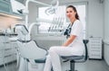 Portraait of sincerely smiling young dentist woman dressed white medical scrubs uniform sitting in modern dental clinic next to