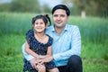 Portrai of young indian farther with his cute little daughter sitting outdoor at green agriculture field background, looking at