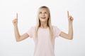 Portrait of adorable amazed child with blond hair in pink t-shirt, raising index fingers, pointing and looking up with