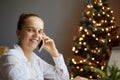 Portrait of adorable Caucasian smiling woman wearing white shirt sitting on table near christmas tree and talking on cell phone, Royalty Free Stock Photo