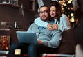 Portrait of an adorable couple with laptop - charming woman in Santa hat hugging her man and looking at camera. Royalty Free Stock Photo