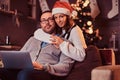 Portrait of an adorable couple with laptop - charming woman in Santa hat hugging her man and looking at camera. Royalty Free Stock Photo