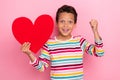 Portrait of adorable ecstatic boy with curly haircut dressed striped long sleeve hold red heart fist up isolated on pink Royalty Free Stock Photo