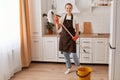 Portrait of adorable hardworking female wearing brown apron and rubber gloves making fast domestic work in white kitchen, holding Royalty Free Stock Photo