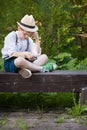 Portrait of adorable kid boy in a straw hat sitting on a wooden bench and reading book in the park. Child reading in the garden on Royalty Free Stock Photo