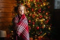 Portrait of adorable little blonde curly child girl wrapped in plaid looking at camera standing by decorated Christmas Royalty Free Stock Photo