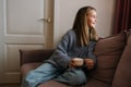 Portrait of adorable little girl holding in hands mug with hot cocoa sitting on sofa by illuminated Christmas tree with Royalty Free Stock Photo