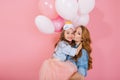 Portrait of adorable long-haired birthday girl in sleep mask holding colorful balloons and embracing her stylish mom