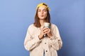 Portrait of adorable positive teenager girl wearing baseball cap and jacket, holding thermos with hot beverage, sending air